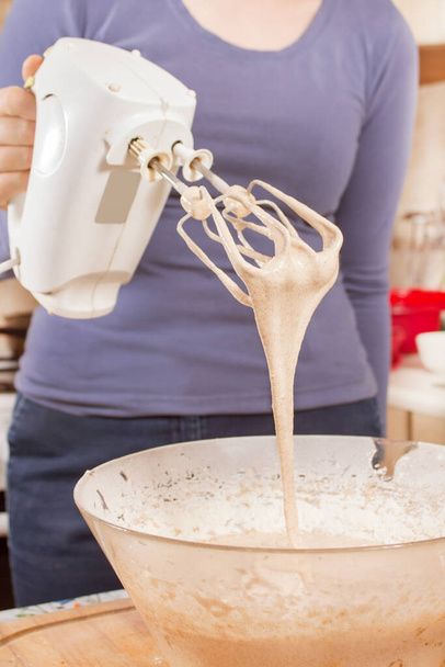 A nő egy mixert tart a kezében. A tészta a keverőlapátokból folyik a tálba. Egy nő sütésének előkészítése. - Fotó, kép
