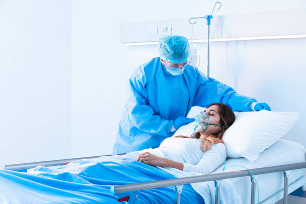 Jovem em pressão positiva de oxigênio no hospital na enfermaria durante a pandemia de Covid-19 sendo assistida por uma enfermeira em EPI
 - Foto, Imagem