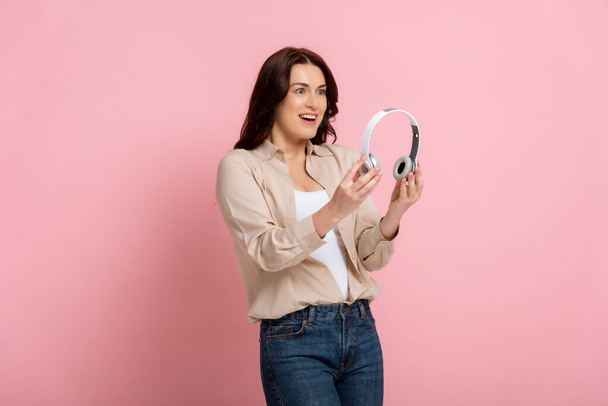Positive brunette woman holding headphones on pink background - Foto, Imagen