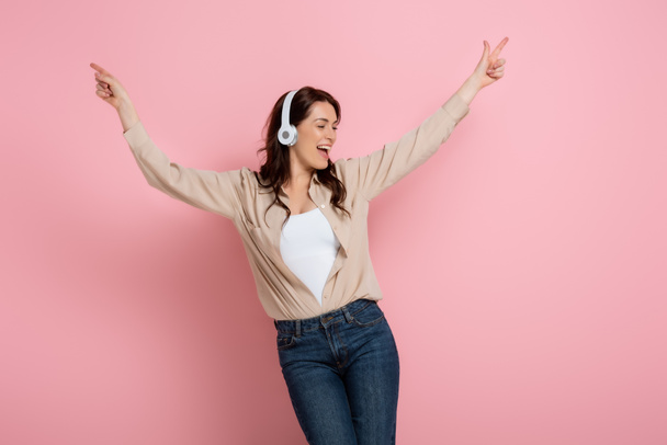 Cheerful woman pointing with fingers while listening music in headphones on pink background - Foto, Bild