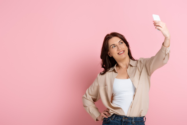 Mujer morena sonriente posando mientras toma selfie en el teléfono inteligente sobre fondo rosa
 - Foto, Imagen