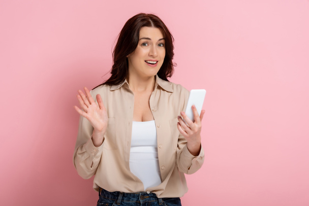 Smiling woman looking at camera while having video call on smartphone on pink background - Photo, Image