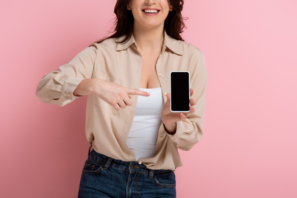 Oříznutý pohled na usmívající se žena ukazuje prstem na smartphone na růžovém pozadí - Fotografie, Obrázek