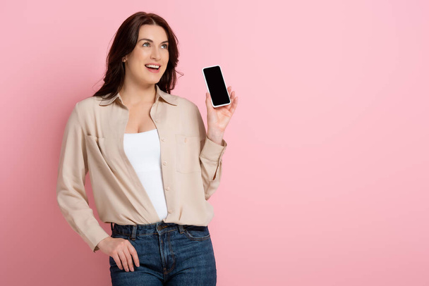 Attractive brunette woman smiling while showing smartphone with blank screen on pink background - Foto, Imagem