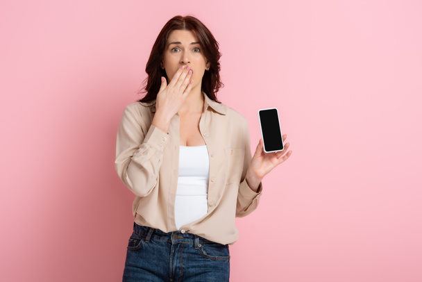 Szokująca kobieta zakrywająca usta ręką i pokazująca smartfon na różowym tle, koncepcja ciała pozytywnego  - Zdjęcie, obraz