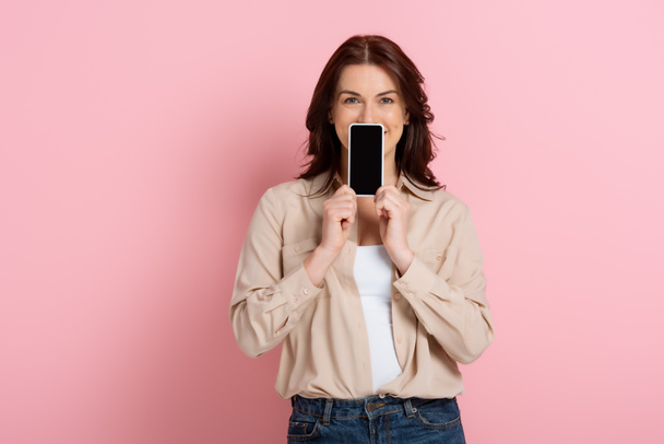 Mujer morena mostrando teléfono inteligente con pantalla en blanco cerca de la cara sobre fondo rosa
 - Foto, imagen