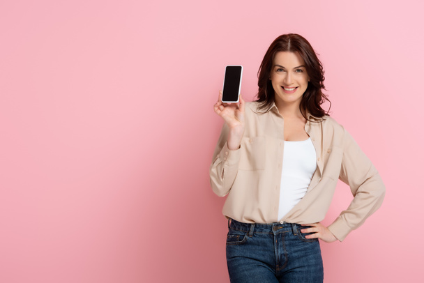 Mooie vrouw glimlachen op camera terwijl het tonen van smartphone met leeg scherm op roze achtergrond - Foto, afbeelding