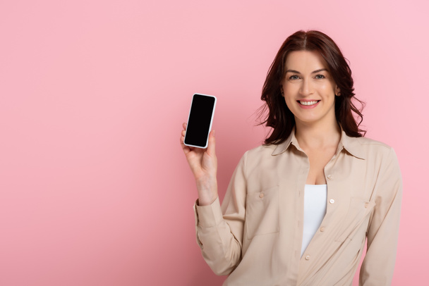 Atraktivní brunetka žena usmívá na kameru, zatímco ukazuje smartphone s prázdnou obrazovkou na růžovém pozadí - Fotografie, Obrázek