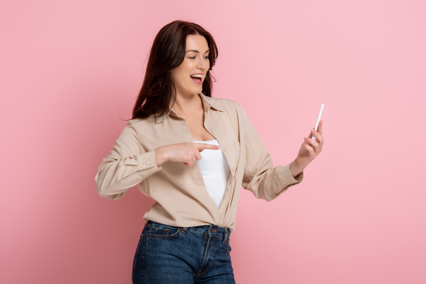 Pozytywna kobieta wskazująca palcem na smartfona na różowym tle - Zdjęcie, obraz