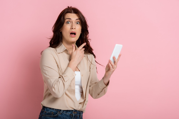 Scared woman looking at camera and pointing with finger while holding smartphone on pink background - Foto, immagini