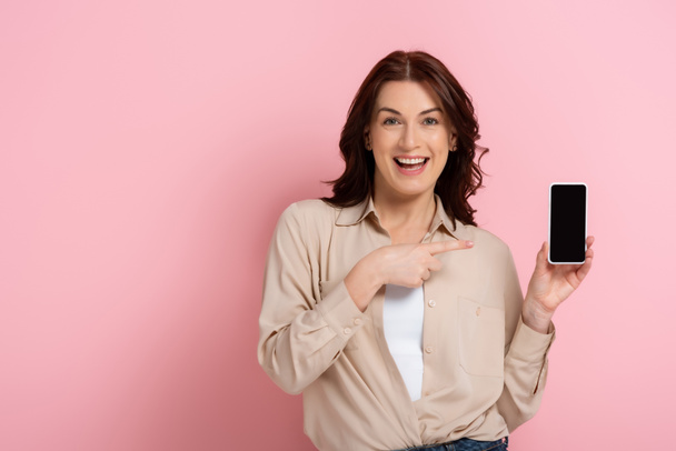 Beautiful woman smiling at camera while pointing with finger at smartphone with blank screen on pink background - Zdjęcie, obraz