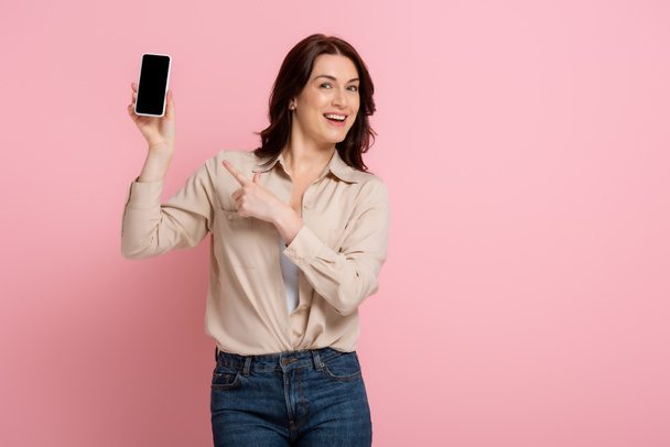 Pozytywna brunetka wskazująca palcem na smartfona z czystym ekranem na różowym tle - Zdjęcie, obraz