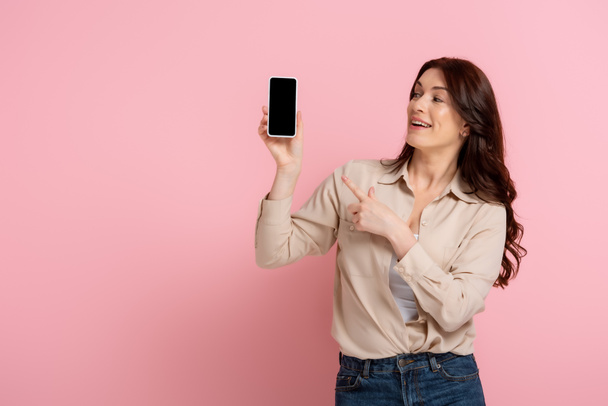 Happy brunette woman pointing with finger at smartphone with blank screen on pink background - Fotoğraf, Görsel