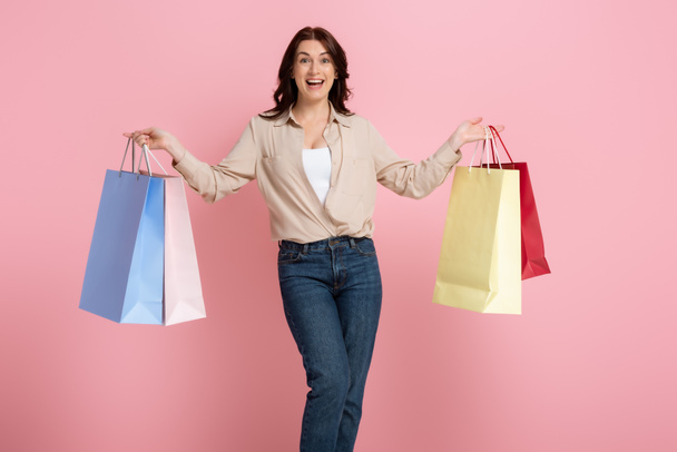 Pozytywna kobieta trzymająca torby na zakupy i patrząca w kamerę odizolowana na różowo, koncepcja body positive  - Zdjęcie, obraz