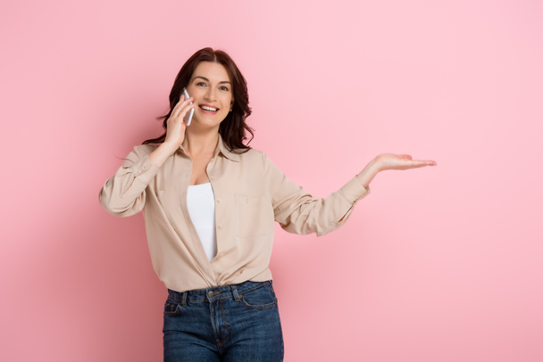 Beautiful smiling woman pointing with hand while talking on smartphone on pink background - Fotó, kép