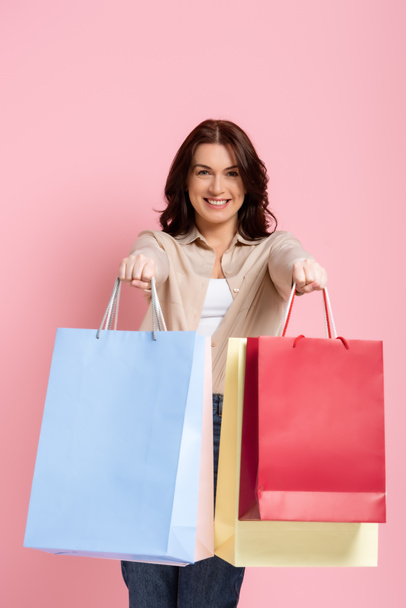 Happy brunette woman smiling at camera and showing shopping bags on pink background - Fotografie, Obrázek