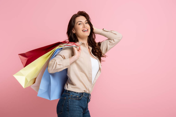 Beautiful brunette woman smiling at camera while holding colorful shopping bags on pink background - Fotografie, Obrázek