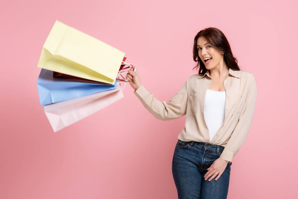 Mulher alegre atraente sorrindo para a câmera enquanto segurando sacos de compras isolados em rosa, conceito de corpo positivo
  - Foto, Imagem