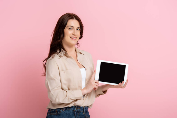 Brunette woman smiling at camera while holding digital tablet on pink background - Foto, immagini