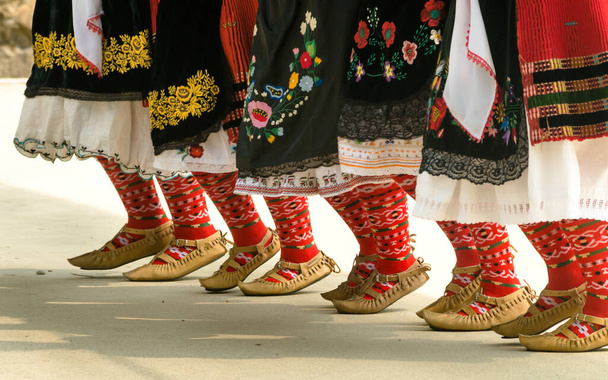 Folclore búlgaro. Chicas bailando danza folclórica. La gente con trajes tradicionales baila danzas folclóricas búlgaras. Primer plano de las piernas femeninas con zapatos, calcetines y trajes tradicionales para bailes folclóricos búlgaros
. - Foto, Imagen