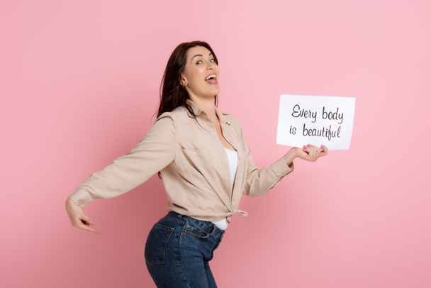 Happy woman pointing with finger on butt while holding card with every body is beautiful lettering on pink background - Fotó, kép