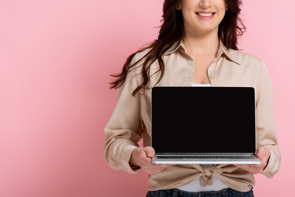 ピンクの背景に空白の画面とノートパソコンを示す笑顔の女性の作物のビュー - 写真・画像