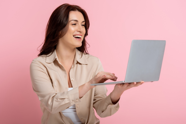 Attractive brunette woman smiling while using laptop on pink background - Foto, Bild