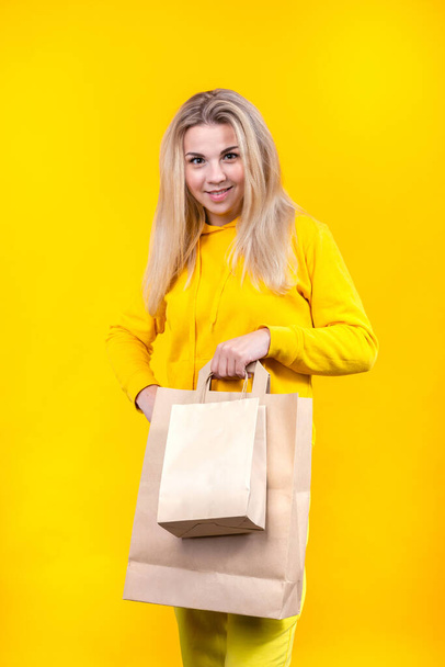 Портрет молодой белокурой блондинки с бумажными эко-пакетами в жёлтом спортивном костюме, изолированной на жёлтом фоне, улыбающейся, смотрящей в камеру
. - Фото, изображение