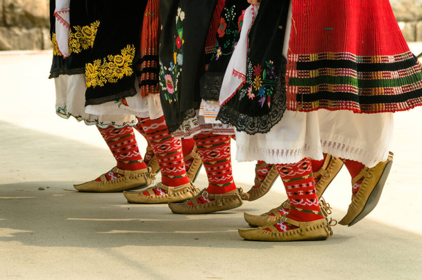 ブルガリアの民間伝承。民俗舞踊を踊る女の子。民族衣装を着た人々がブルガリアの民族舞踊を踊る。ブルガリアの民俗舞踊のための伝統的な靴、靴下、衣装を持つ女性の足のクローズアップ. - 写真・画像