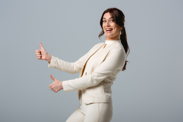 Vista lateral da bela mulher de negócios sorridente mostrando polegares isolados em cinza
  - Foto, Imagem