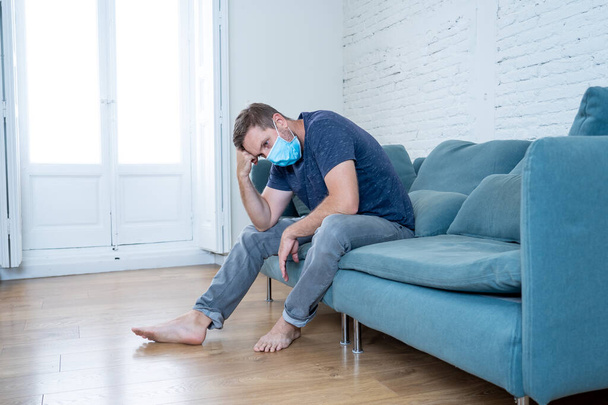 Sad man with protective face mask at home living room couch feeling tired and worried suffering depression amid coronavirus lockdown and social distancing. Mental Health and isolation concept. - Fotoğraf, Görsel