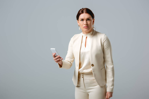Confused businesswoman looking at camera while holding smartphone isolated on grey  - Фото, изображение