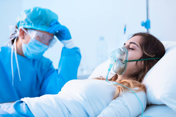 Dramatic side view of a young female patient breathing assisted by a respiratory tube and an exhausted dedicated doctor in the intensive care unit of a hospital - 写真・画像