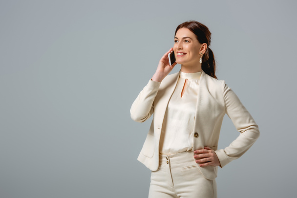 Mujer atractiva en ropa formal sonriendo mientras habla en un teléfono inteligente aislado en gris
  - Foto, imagen