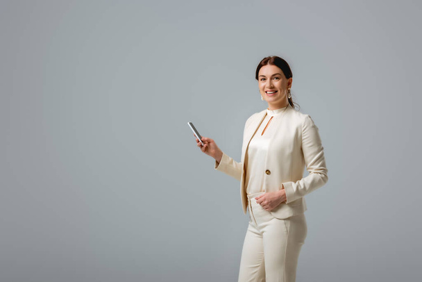 Beautiful woman in formal wear smiling at camera while holding smartphone isolated on grey  - Foto, Imagem