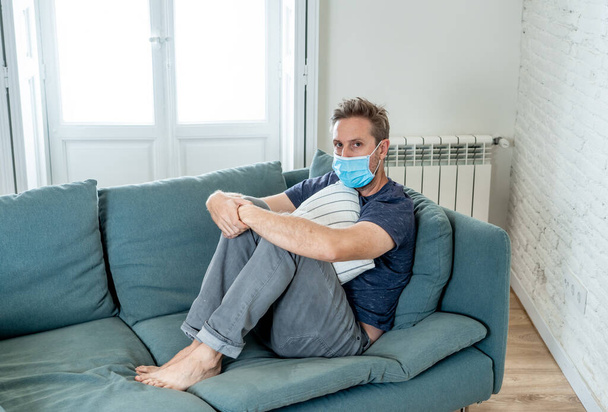Sad man with protective face mask at home living room couch feeling tired and worried suffering depression amid coronavirus lockdown and social distancing. Mental Health and isolation concept. - 写真・画像