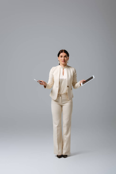 Brunette businesswoman looking at camera while holding digital devices on grey background - Zdjęcie, obraz