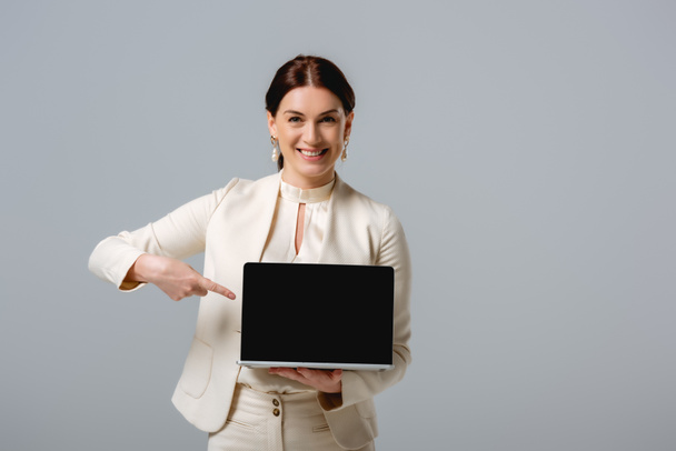 Piękna bizneswoman wskazująca palcem na laptopa i uśmiechnięta do kamery odizolowana na szaro - Zdjęcie, obraz