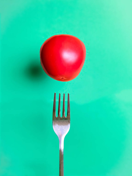Ισορροπώντας την ντομάτα πάνω από ένα πιρούνι σε τυρκουάζ φόντο. Υγιεινή διατροφική έννοια. Πρότυπο μαγειρέματος. - Φωτογραφία, εικόνα