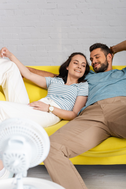 Focus selettivo della giovane donna sdraiata accanto al fidanzato sorridente sul divano vicino al ventilatore elettrico  - Foto, immagini