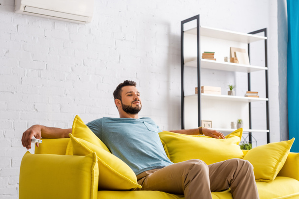 Schöner Mann hält Fernbedienung der Klimaanlage auf Couch  - Foto, Bild
