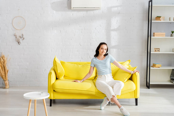 Mulher bonita olhando para a câmera enquanto sentado no sofá perto do controlador remoto de ar condicionado na mesa de café
  - Foto, Imagem