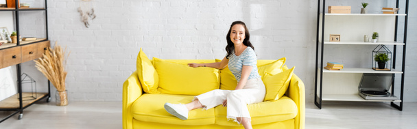 Panoramic shot of young woman smiling at camera while sitting on yellow sofa at home  - Valokuva, kuva
