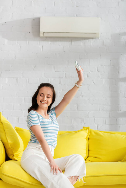 リビングでソファの上にエアコンのリモコンを持っている笑顔の女性 - 写真・画像