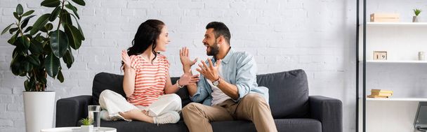 Panoramic crop of cheerful couple looking at each other on couch at home  - Foto, Imagen