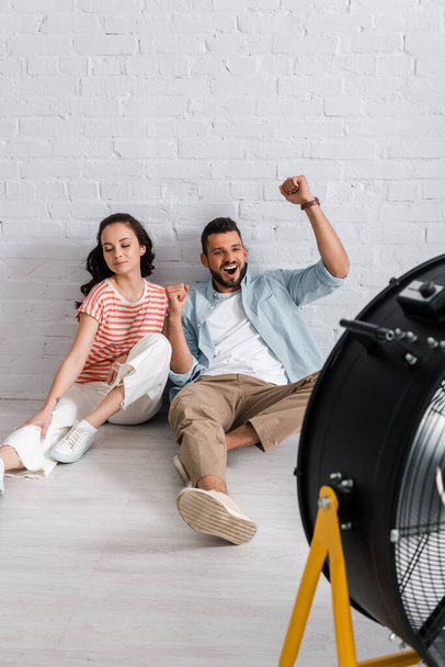 foyer sélectif de l'homme joyeux montrant oui geste près de la petite amie et ventilateur électrique sur le sol  - Photo, image