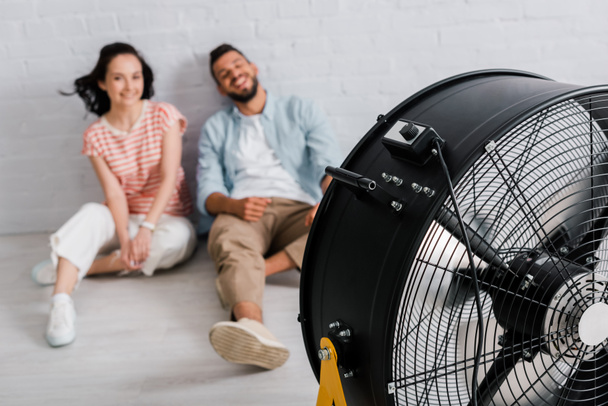 Selective focus of electric fan near smiling couple sitting on floor at home  - Valokuva, kuva