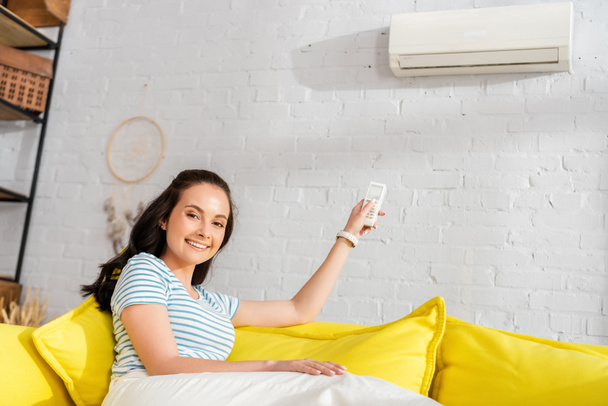 Young woman smiling at camera while switching air conditioner with remote controller on sofa  - Foto, imagen
