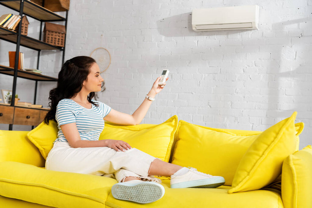 Schöne lächelnde Frau schaltet Klimaanlage, während sie zu Hause auf gelbem Sofa sitzt  - Foto, Bild