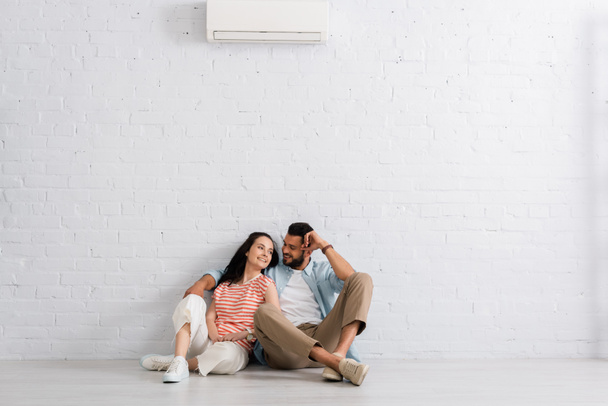 Hezký muž objímající krásnou přítelkyni, zatímco sedí pod klimatizací na podlaze doma  - Fotografie, Obrázek